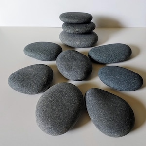 May include: A collection of smooth, gray river stones. Some are stacked, while others are arranged around the stack. The stones vary in size and shape, with a natural, weathered appearance. The stones are a neutral gray color.