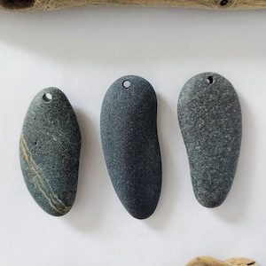 May include: Three gray, smooth river stones with drilled holes for hanging. The stones vary in shape and texture, with one showing a stripe of yellow and tan. Driftwood pieces frame the stones on a white background.