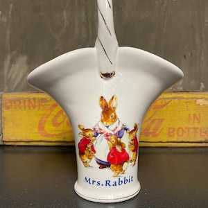 May include: White ceramic basket with a gold handle and a depiction of Mrs. Rabbit from Beatrix Potter's children's book, "The Tale of Peter Rabbit." The basket features Mrs. Rabbit surrounded by three bunnies, all wearing red jackets.