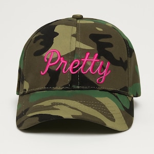 May include: A camouflage baseball cap with the word "Pretty" embroidered in bright pink cursive across the front. The cap features a curved brim and a button on top. The camouflage pattern includes shades of green, brown, and black.