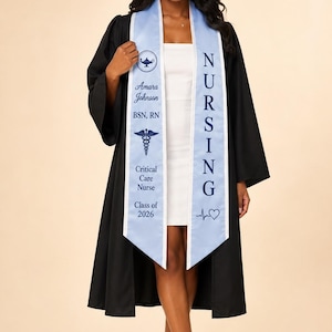 Puede incluir: Una toga de graduación negra sobre un vestido blanco, combinada con una estola de graduación azul claro. La estola presenta el texto 'NURSING', 'Amara Johnson', 'BSN, RN', 'Critical Care Nurse', 'Class of 2026', un icono de lámpara, un caduceo y un corazón.