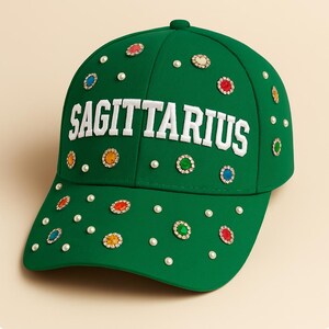 May include: A green baseball cap with the word "SAGITTARIUS" in white block letters. The cap is adorned with colorful jewel-like embellishments and small white pearls.