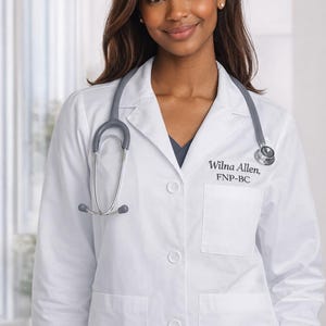 May include: A white medical coat with a stethoscope. The coat has pockets and buttons down the front. Text on the coat reads "Wilna Allen, FNP-BC". The person is smiling.