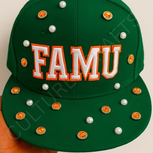 May include: A green snapback hat with the word "FAMU" in white letters with orange trim. The hat is decorated with faux pearls and orange-colored jewels. The hat has a flat brim.