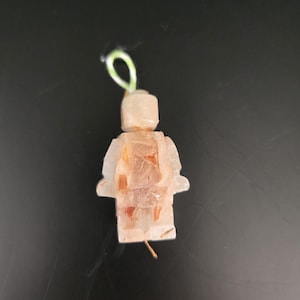 May include: A clear, Lego-shaped ornament with embedded orange and brown elements. The ornament has a green loop at the top for hanging. The figure is a classic toy design, suitable for holiday or everyday decor.