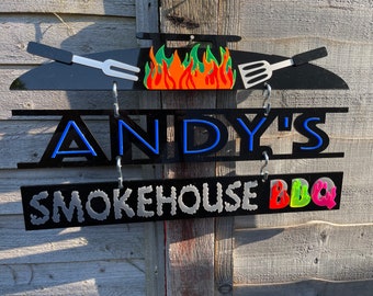 May include: Black acrylic sign with the text "ANDY'S SMOKEHOUSE BBQ". The sign features a grill design with flames and utensils. The text is in various colors, including blue, silver, red, and green.