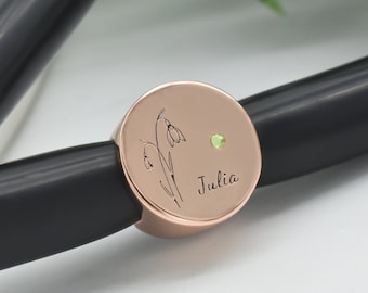 May include: Rose gold stethoscope charm with a green gemstone and an engraved image of a flower with the name "Julia" inscribed.