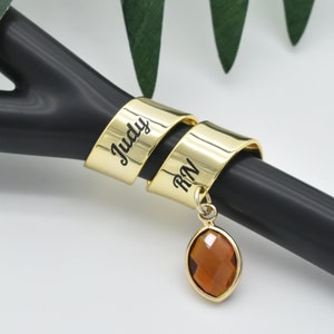 May include: Gold stethoscope charm engraved with the name "Judy RN". The charm features a brown gemstone pendant.