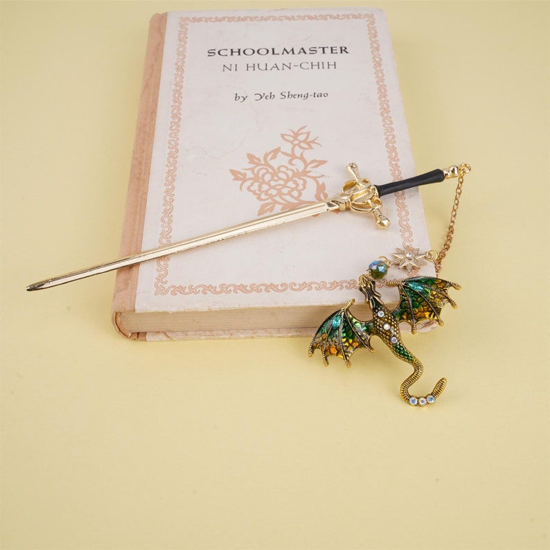 May include: Gold dragon hair stick with green and gold enamel and clear rhinestones. The dragon is attached to a gold chain with a sword charm. The hair stick is resting on a book titled "Schoolmaster Ni Huan-Chih by Yeh Sheng-tao".
