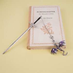 May include: A silver sword bookmark with a purple dragon charm hanging from a chain. The bookmark is resting on a book titled "Schoolmaster Ni Huan-Chih" by Yeh Sheng-tao.