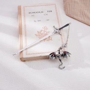 May include: Silver bookmark with a sword design and a dragon pendant with red gem accents. The bookmark is resting on a closed book with the text "SCHOOLMASTER" on the cover.