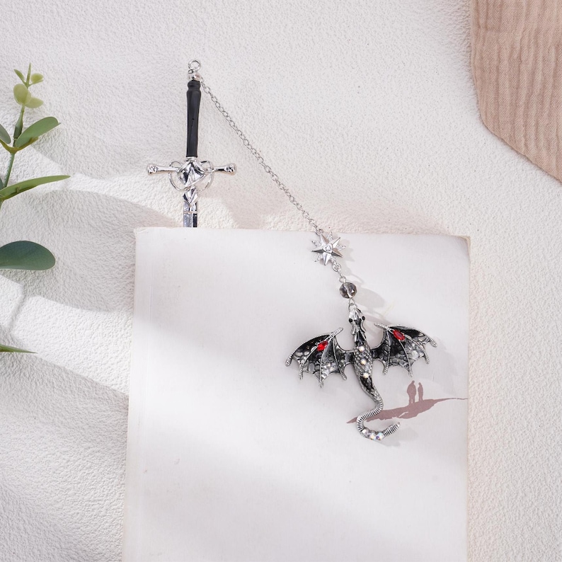 May include: A silver sword and dragon bookmark with a chain connecting them. The dragon is black with red eyes and is detailed with silver accents. The sword has a silver handle and a black blade.