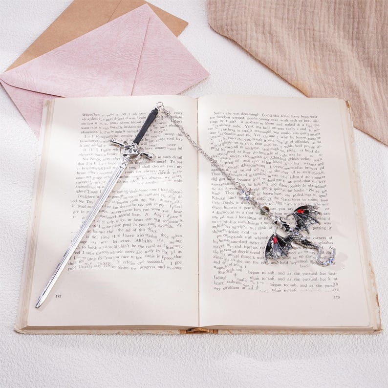 Peut inclure: Deux pendentifs de marque-page en argent, l'un en forme d'&eacute;p&eacute;e et l'autre en forme de dragon. Le dragon a des yeux rouges et des ailes noires. Les deux pendentifs sont sur une cha&icirc;ne et sont plac&eacute;s sur un livre ouvert.