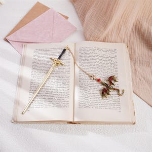 Peut inclure: Signet de livre en or avec un motif d'&eacute;p&eacute;e &agrave; une extr&eacute;mit&eacute; et un motif de dragon rouge et vert &agrave; l'autre extr&eacute;mit&eacute;. Le signet est pos&eacute; ouvert sur un livre.