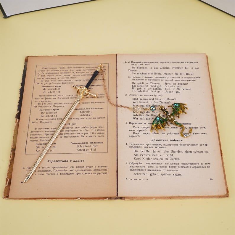 May include: A gold-tone bookmark with a green and gold dragon charm. The bookmark is attached to a chain and has a sword-shaped handle. The bookmark is open to a page of a German language textbook.