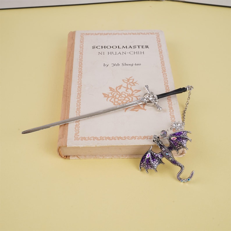 May include: A silver sword bookmark with a purple dragon charm. The bookmark is resting on a book titled "Schoolmaster Ni Huan-Chih by Yeh Sheng-tao".