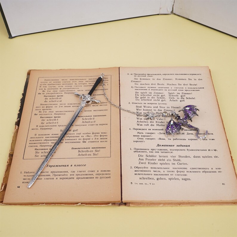 May include: A silver sword bookmark with a purple and silver dragon charm. The bookmark is open on a book with German text.
