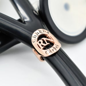 Pode incluir: Um amuleto de estetoscópio de ouro rosa com as letras "RN" em forma de coração e o nome "Chacha Carlei" gravado em torno do coração.