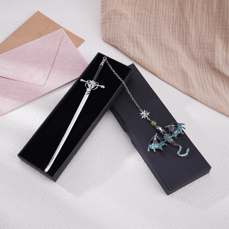 May include: Two black velvet boxes with silver sword and green dragon bookmarks. The sword bookmark has a silver handle and a black blade. The dragon bookmark has green and silver wings and a silver chain.