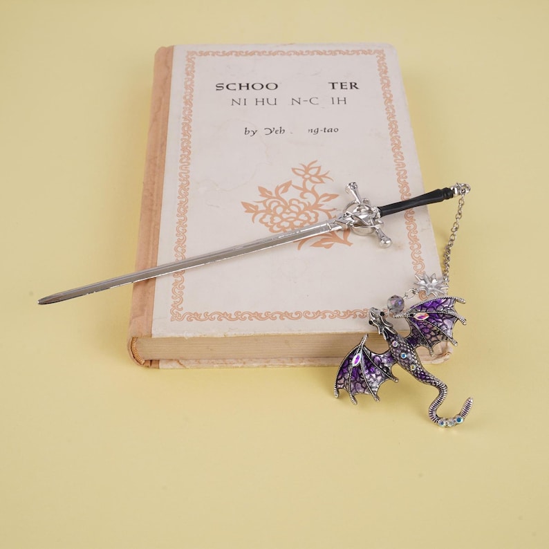 Peut inclure: Un marque-page en forme de dragon argent&eacute; avec des accents en &eacute;mail violet et blanc et un marque-page en forme d'&eacute;p&eacute;e argent&eacute;e. Le marque-page en forme de dragon est dot&eacute; d'une cha&icirc;ne. Les marque-pages sont pos&eacute;s sur un livre ferm&eacute; intitul&eacute; "Schoolmaster" par "Yeh. ng-tao".