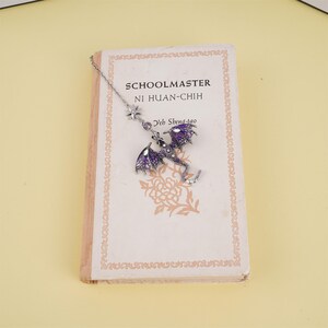May include: A silver bookmark with a purple dragon charm and a star charm. The bookmark is hanging from a book titled "Schoolmaster Ni Huan-Chih Yeh Shena-tao".