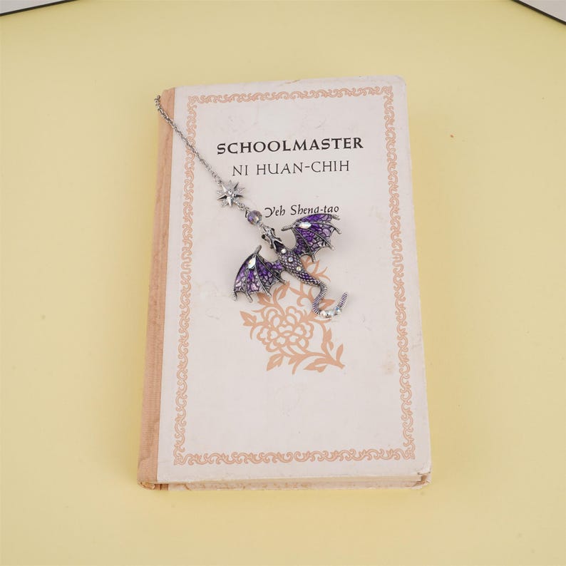 May include: A silver bookmark with a purple dragon charm and a star charm. The bookmark is hanging from a book titled "Schoolmaster Ni Huan-Chih Yeh Shena-tao".