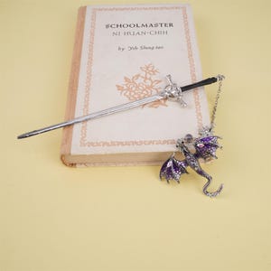 May include: A silver bookmark with a dragon design. The dragon has purple wings and a silver body with clear rhinestones. The bookmark also includes a silver sword with a black handle.