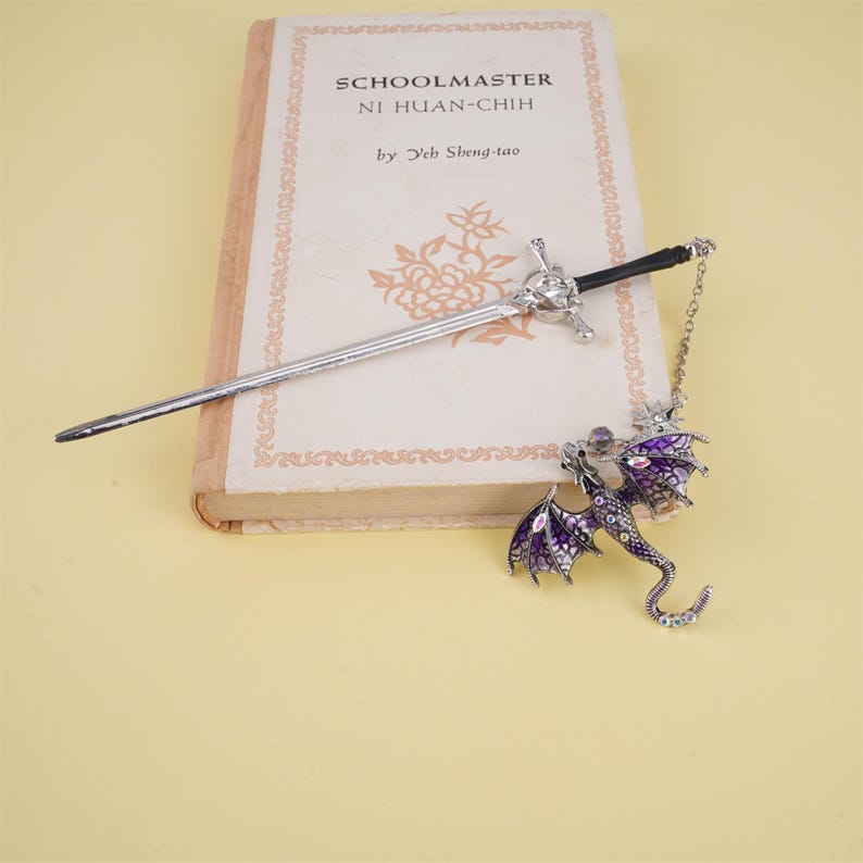 May include: A silver bookmark with a dragon design. The dragon has purple wings and a silver body with clear rhinestones. The bookmark also includes a silver sword with a black handle.