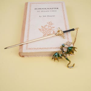 Peut inclure: Un marque-page en forme de dragon dor&eacute; avec un &eacute;mail vert et jaune et des strass transparents. Le dragon tient une pierre pr&eacute;cieuse transparente iris&eacute;e. Le marque-page est attach&eacute; &agrave; un b&acirc;ton de cheveux en forme d'&eacute;p&eacute;e dor&eacute;e.