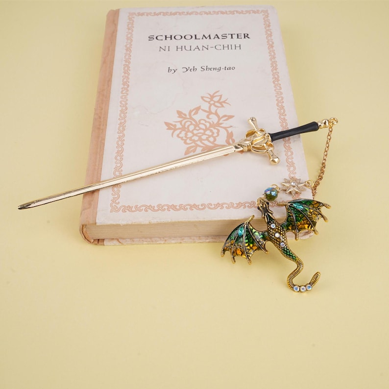 Peut inclure: Un marque-page en forme de dragon dor&eacute; avec un &eacute;mail vert et jaune et des strass transparents. Le dragon tient une pierre pr&eacute;cieuse transparente iris&eacute;e. Le marque-page est attach&eacute; &agrave; un b&acirc;ton de cheveux en forme d'&eacute;p&eacute;e dor&eacute;e.