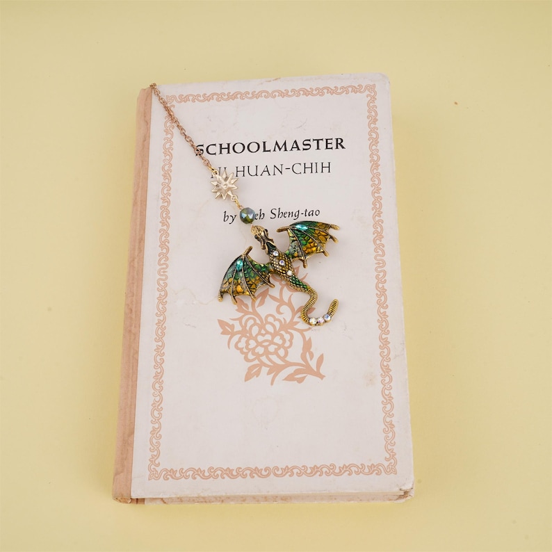 May include: A gold chain bookmark with a green and gold dragon charm and a small star charm. The bookmark is on top of a book titled "Schoolmaster" by "Yeh Sheng-tao".