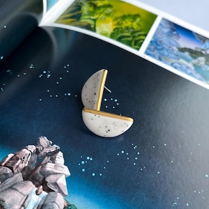 May include: A pair of half-moon earrings. The earrings are white with black speckles and gold trim. The post is gold. The earrings are on a dark blue surface with white dots.
