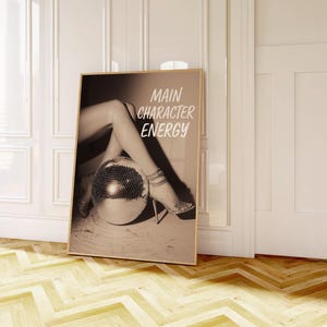 May include: Sepia-toned framed print with the text "MAIN CHARACTER ENERGY." The image features legs and high heels resting on a disco ball. The print is leaning against a white wall in a room with a wooden floor.