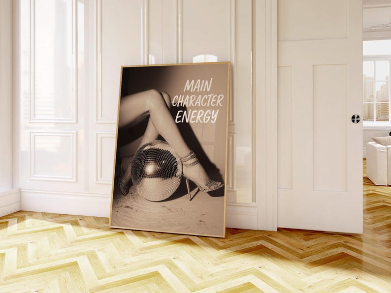 May include: Sepia-toned framed print with the text "MAIN CHARACTER ENERGY." The image features legs and high heels resting on a disco ball. The print is leaning against a white wall in a room with a wooden floor.
