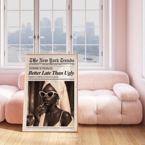 May include: A framed sepia-toned print titled "The New York Trends" with the headline "Better Late Than Ugly." The image features a woman wearing sunglasses and a towel, holding a lipstick. The print is displayed in a pink room with a pink sofa and a window.