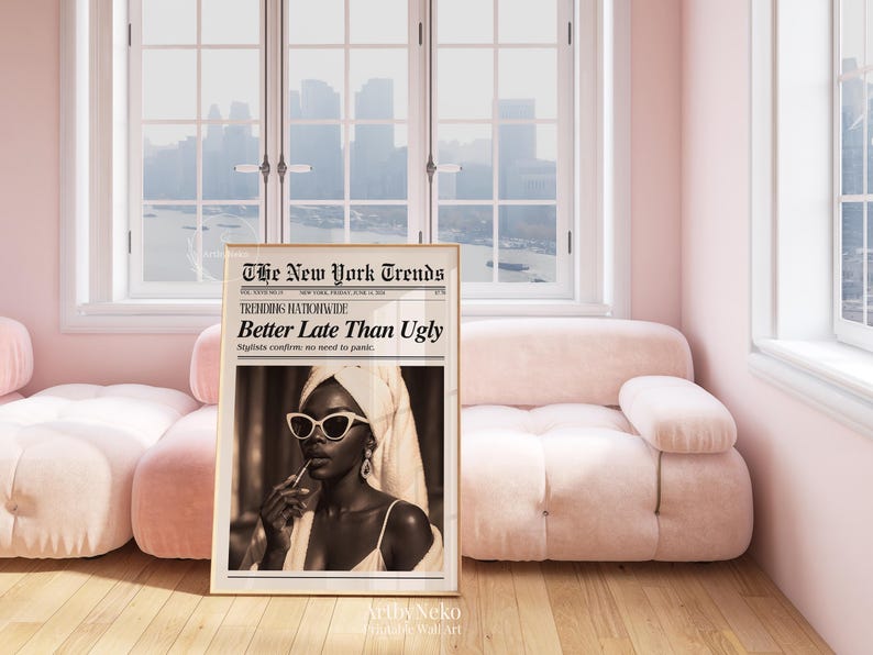 May include: A framed sepia-toned print titled "The New York Trends" with the headline "Better Late Than Ugly." The image features a woman wearing sunglasses and a towel, holding a lipstick. The print is displayed in a pink room with a pink sofa and a window.