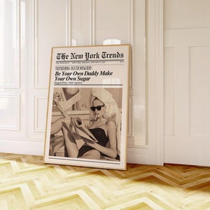 May include: A framed sepia-toned print of a newspaper titled "The New York Trends" with the headline "Be Your Own Daddy Make Your Own Sugar." The image within shows a woman in sunglasses and a towel, holding a newspaper.