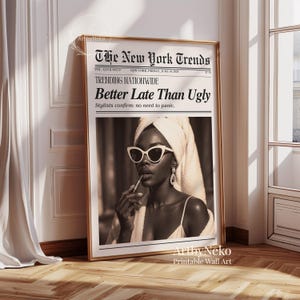 May include: A framed print featuring a sepia-toned image of a woman wearing sunglasses and applying lipstick. The print is designed to look like a newspaper, with the headline "Better Late Than Ugly" and "The New York Trends" at the top. The frame is gold.