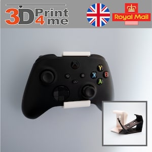 May include: A white 3D printed wall mount for an Xbox controller. The mount is attached to a black Xbox controller. The controller is mounted on a white wall. The image also shows a close-up of the mount.