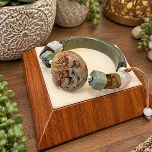 Puede incluir: Una pulsera que presenta una pieza central grande, redonda y de piedra marrón tallada con motivos florales, flanqueada por cuentas más pequeñas verdes, marrones y translúcidas. La banda principal es un segmento de piedra verde oscuro liso y curvo.