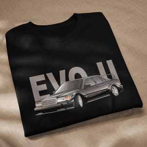 Op de afbeelding: Zwart T-shirt met een zilveren illustratie van een klassieke zwarte auto met een achterspoiler. De tekst "EVO II" is in grote zilveren letters boven de auto gedrukt. Het shirt is gemaakt van zacht materiaal.