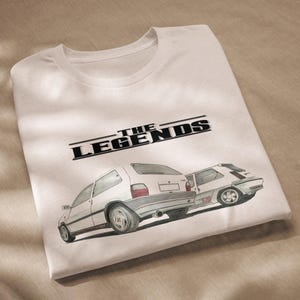 Può includere: Maglietta bianca con una grafica nera di due auto bianche e il testo "THE LEGENDS" sopra.