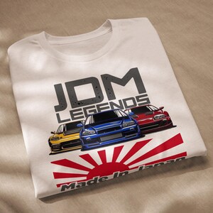 May include: White t-shirt with a graphic design featuring the text "JDM LEGENDS" in black and grey, three cartoon cars in yellow, blue, and red, and a red and white rising sun with the text "Made in Japan".