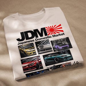 Może przedstawiać: Biała koszulka z czarno-białym nadrukiem z tekstem "JDM Japanese Domestic Market" z czerwono-białym wzorem wschodzącego słońca. Nadruk zawiera również ilustracje różnych japońskich samochodów.