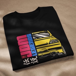 Può includere: Maglietta nera con un'illustrazione di un'auto gialla e il testo "Legends" in blu, "Sounds" in rosa e "JDM" in rosso. Il testo giapponese si traduce in "Fast and Furious".