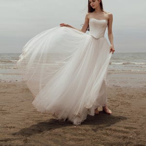 May include: A strapless white wedding dress with a corset bodice and a flowing tulle skirt. The dress is worn on a beach with the ocean in the background. The dress is floor-length and has a delicate, ethereal quality.
