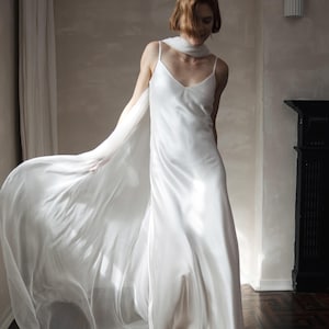 May include: A white satin slip dress with spaghetti straps and a flowing, sheer scarf. The dress is floor-length and paired with white pointed-toe heels. The image is set in a room with a dark fireplace and hardwood floors.