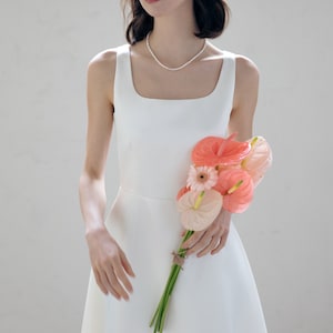 May include: A woman wearing a white sleeveless dress with a pearl necklace and a white headband with pearl accents. She is holding a bouquet of pink and white flowers.