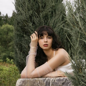 May include: A woman with long, dark curly hair is wearing a white sleeveless top and sheer, light pink gloves. She is leaning on a large, gray rock with a green leafy background.