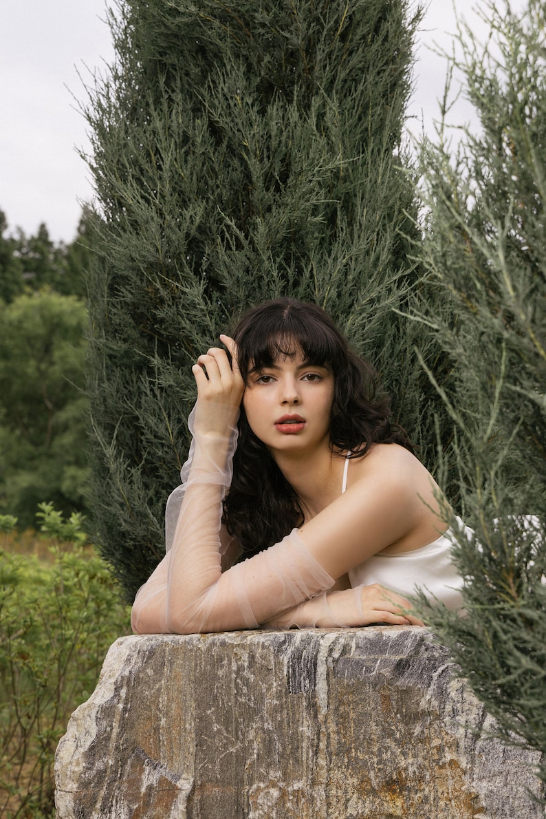 May include: A woman with long, dark curly hair is wearing a white sleeveless top and sheer, light pink gloves. She is leaning on a large, gray rock with a green leafy background.